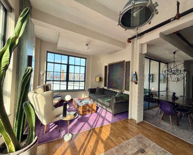 The image shows a spacious, modern living area with large windows, featuring a mix of furniture including a couch, armchairs, and a coffee table on a purple rug.