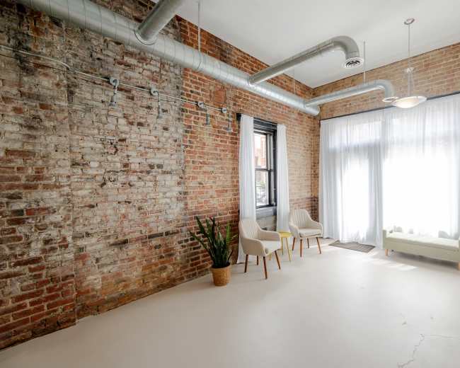A spacious room with exposed brick walls, two chairs, a small couch, and large windows covered by sheer curtains.