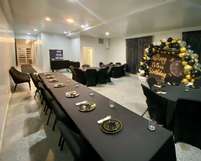 The image shows a decorated party room with black tables set with dinnerware and a large balloon arch featuring "Happy 55th Birthday" in gold and black.