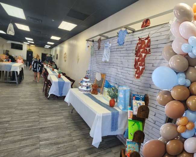 The image shows a decorated event space with tables covered in white tablecloths, a dessert table featuring a cake and gifts, and a colorful balloon arrangement on one side.