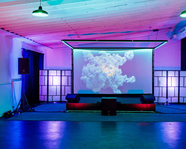 A performance stage features a large screen displaying visual effects, flanked by speakers and illuminated by colorful lighting in a spacious room.