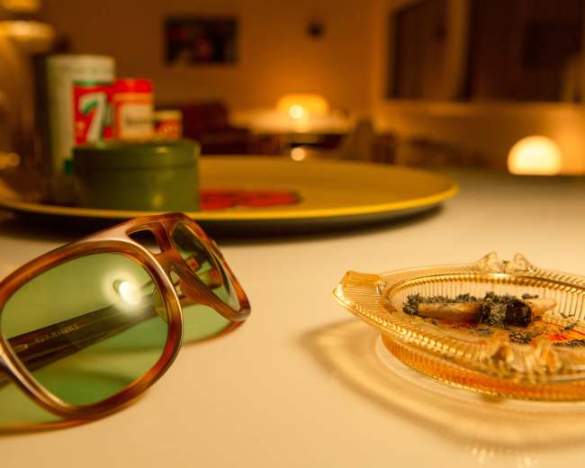 A pair of sunglasses and an ashtray with remnants are placed on a table in a softly lit room.