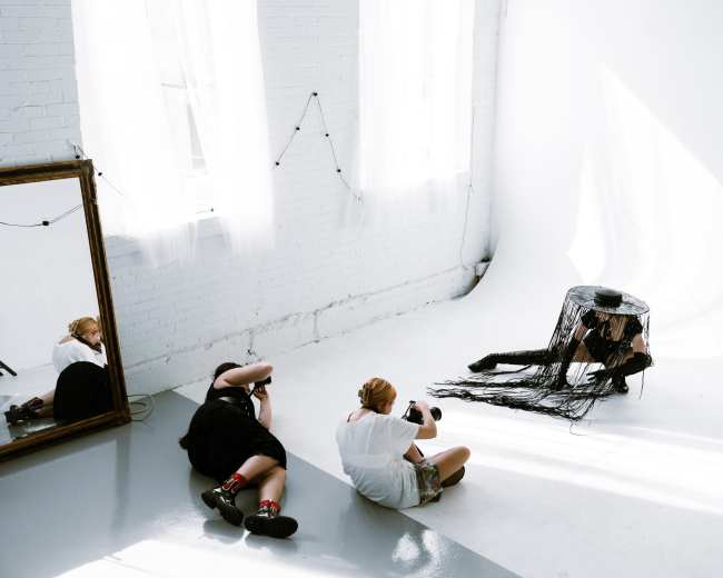 Two photographers capture images of a model posing under a fringed black hat in a bright, minimalist studio with a large mirror.