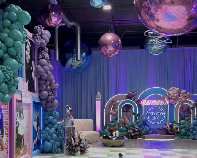 The image depicts a vibrant event space decorated with colorful balloons, floral arrangements, and ambient lighting, featuring a checkered floor and elegant seating.