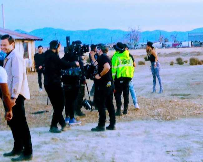 A film crew is gathered in a desert landscape with a performer in the foreground and mountains in the background.