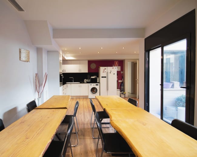 A spacious dining area features long wooden tables and black chairs, leading to a modern kitchen with white cabinetry and appliances.