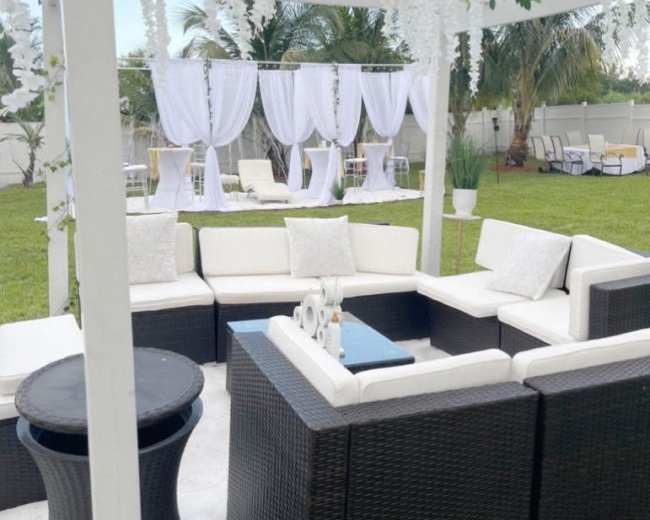 A white pergola adorned with hanging decorations overlooks a spacious outdoor seating area and event setup in a lush green lawn.