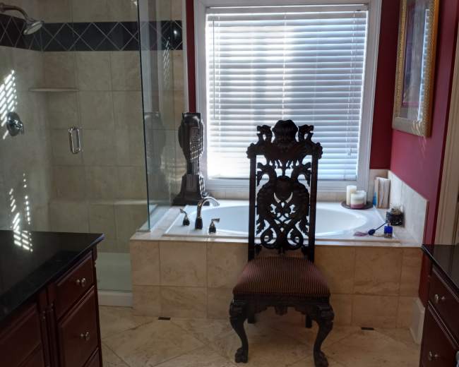 A bathroom features a glass shower enclosure, a tub with a window above it, and a decorative chair near the tub.