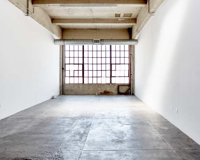 The image shows an empty room with concrete flooring and large windows along one wall, allowing natural light to enter.