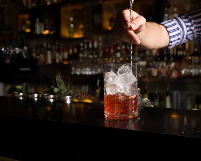 A bartender stirs a cocktail in a glass filled with ice at a bar.