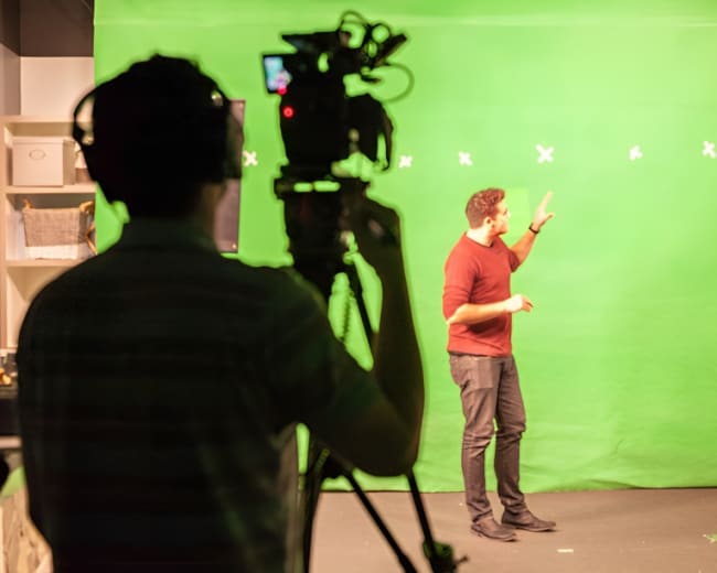 A person gestures in front of a green screen while another individual operates a camera from the foreground.