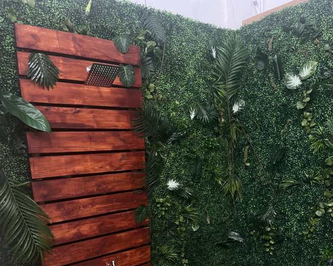 A wooden shower with slatted sides, featuring a shower head and surrounded by green foliage on a grass-like surface.