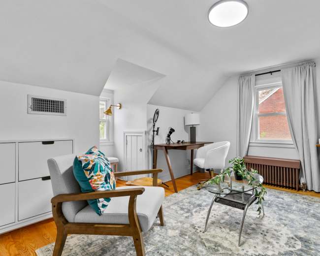 A bright, cozy room featuring a seating area with a gray chair, a small desk, and large windows allowing natural light.