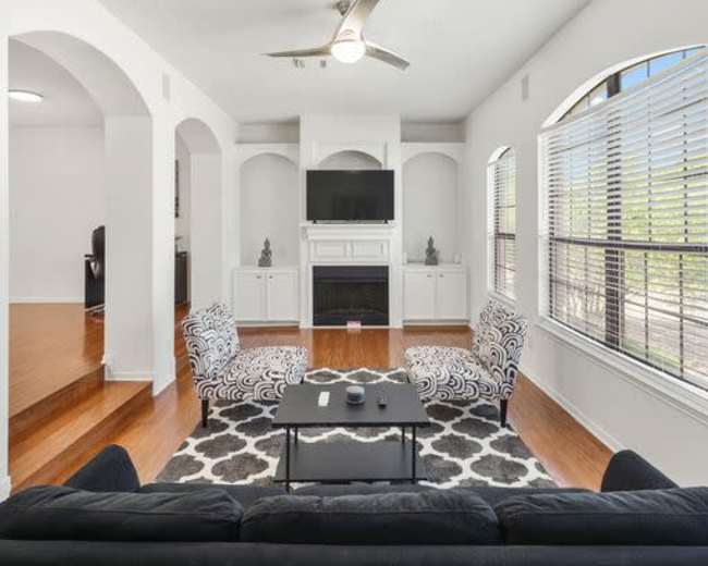 A modern living room features a dark sofa, a coffee table on a patterned rug, and built-in shelving with a television above a fireplace.