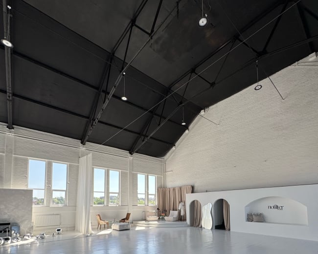 The image shows a spacious, well-lit studio with a high black ceiling, white wooden floors, and minimalistic furniture arranged along the walls.