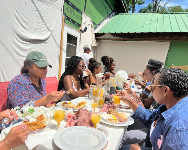 A group of people is seated at a long table outdoors, enjoying a meal that includes various dishes and drinks.