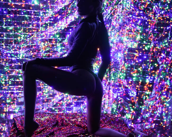 A person poses on a textured surface in front of a vibrant, multicolored sequin backdrop.