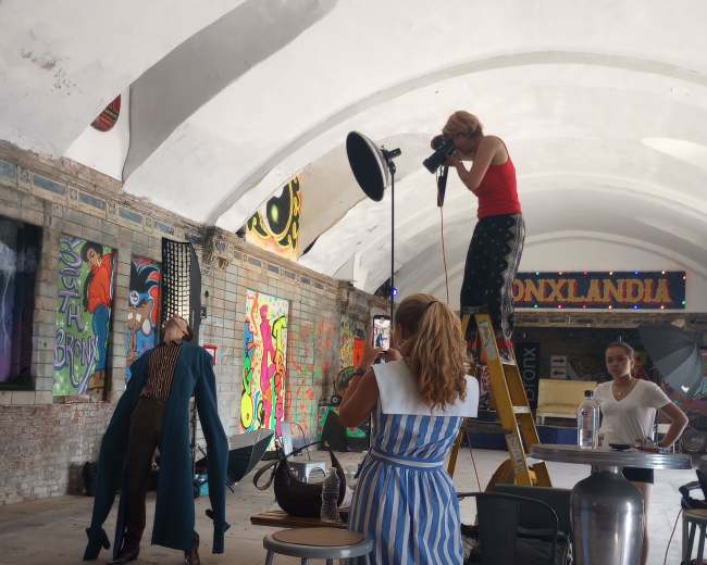 A fashion photoshoot is taking place indoors, featuring a model in a striking outfit and a photographer on a ladder adjusting lights.