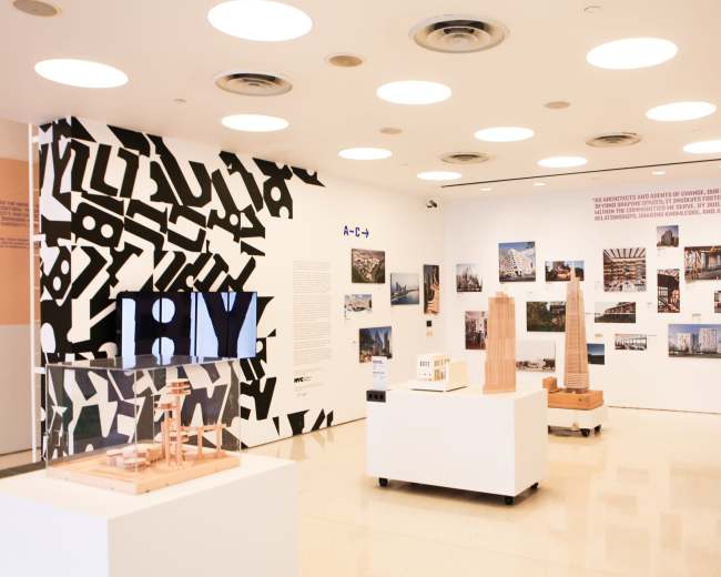 The image shows an exhibition space featuring a large black-and-white patterned wall, various photographs of architectural designs, and display models on white pedestals.