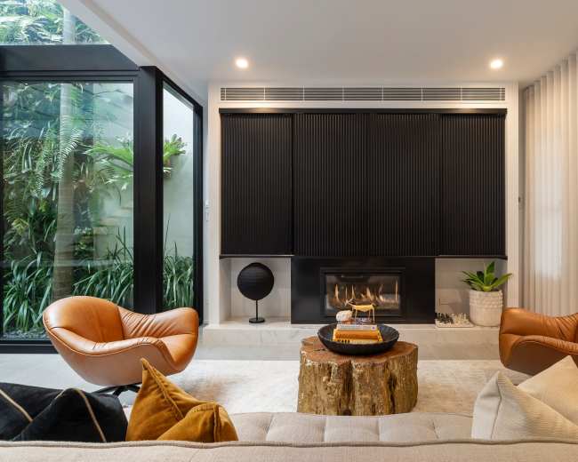 The living room features two orange armchairs, a wooden coffee table, and a fireplace, surrounded by large glass windows and indoor plants.