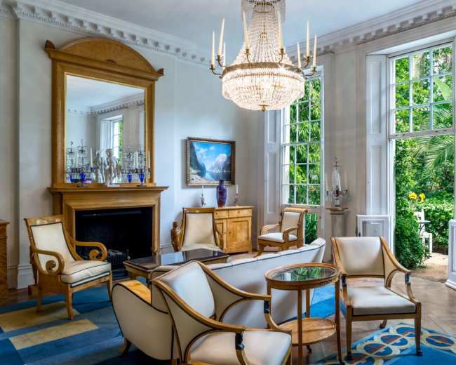 The image depicts a elegantly furnished living room featuring white chairs, a chandelier, a fireplace, and large windows that open to a garden view.