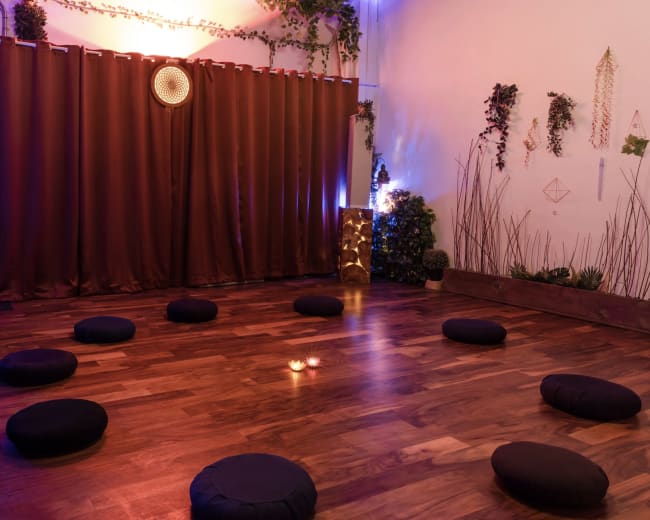 The image depicts a meditation space with round cushions arranged in a circle on polished wooden flooring, illuminated by soft lighting and surrounded by decorative plants and a curtain.