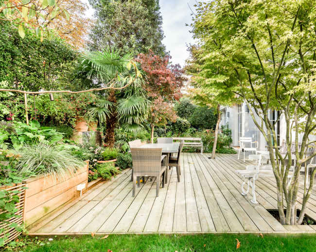 The image depicts a wooden deck surrounded by lush greenery and trees, with a dining table and chairs situated on one side.