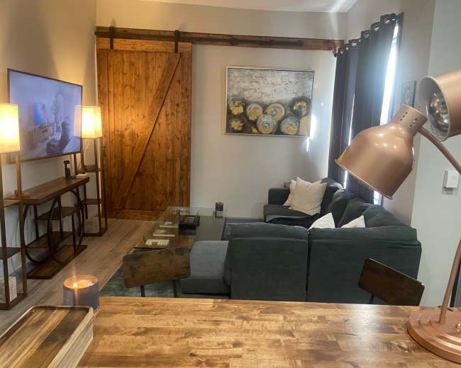 The image shows a modern living room with a dark green sofa, a wooden coffee table, a wall-mounted TV, and a decorative painting, all set against a backdrop of warm wood accents and soft lighting.