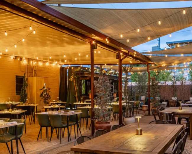 The outdoor dining area features wooden tables, green chairs, and string lights under a shaded canopy.