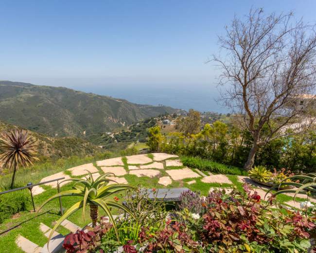 A landscaped garden with various plants and stone pathways overlooking a green hillside and the ocean in the distance.