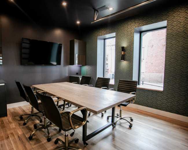 The image features a modern conference room with a large wooden table surrounded by black leather chairs, a wall-mounted television, and two windows.