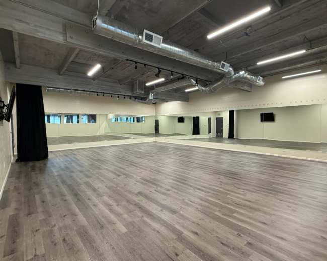 The image shows a spacious dance studio with a wooden floor, large mirrors on one wall, and overhead lighting.