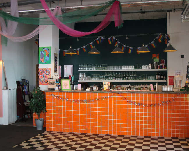 The image shows a vibrant café with an orange tiled counter, glass shelves, and colorful decorations, including hanging lights and a garland.