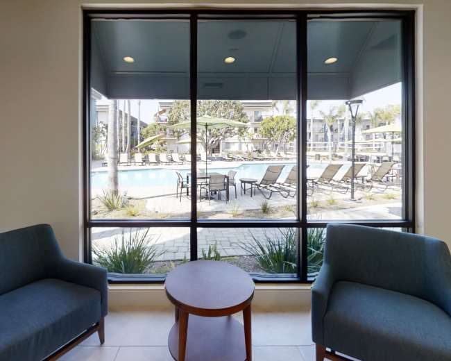 Two blue chairs and a small round table are positioned in front of large windows overlooking a pool area with lounge chairs and greenery.