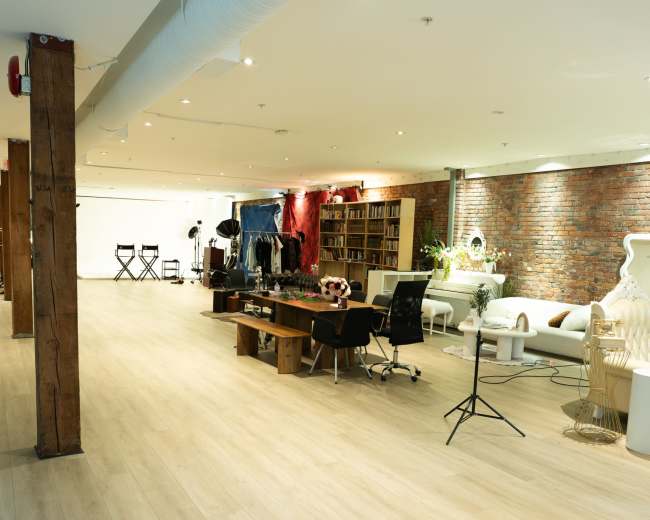 The image shows a spacious interior featuring wooden flooring, exposed brick walls, and a mix of furniture, including a long table, chairs, and shelves filled with books.