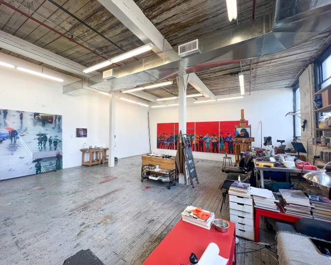 An art studio with wooden floors, a large wall featuring abstract paintings, a wooden work table, and various art supplies scattered around.