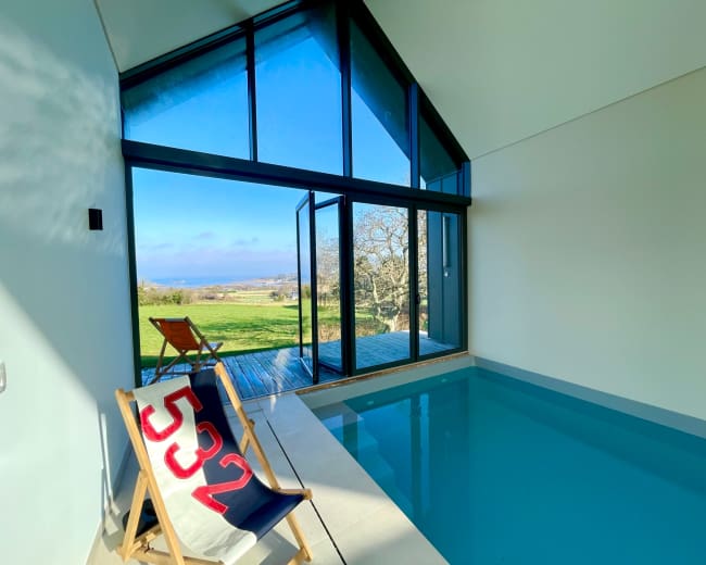 A modern indoor pool area features large glass doors opening to a scenic landscape with a view of trees and the ocean.