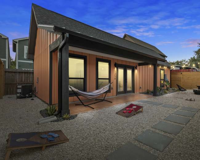 The image shows a modern outdoor area with a gravel patio featuring a hammock, a fire pit, and a pathway lined with stone slabs leading to a house with orange and black siding.