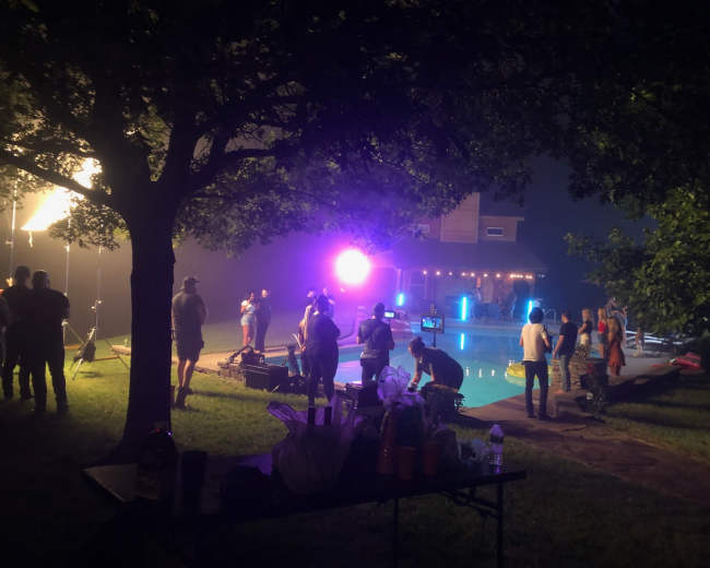 A film crew sets up equipment near a swimming pool illuminated by colorful lights at night.
