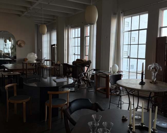 The image shows a spacious interior featuring various tables and chairs, with large windows allowing natural light to fill the room.