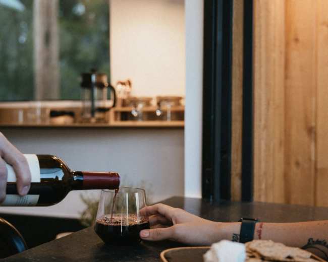 A hand pours red wine into a glass while another hand reaches for a plate of crackers and cheese on a table.