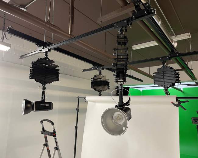 A lighting setup in a studio with multiple adjustable lights mounted on a track above a white backdrop and a green screen.