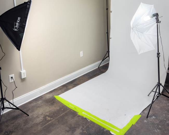 The image shows a photography studio setup with a white backdrop, two lighting equipment stands, and a light reflector positioned on the floor.