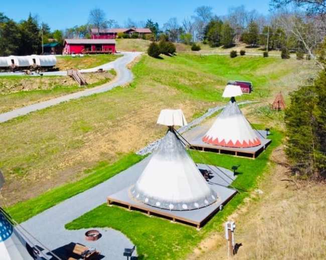 The image shows a landscape with several teepees and a red building in the background, along with a gravel path winding through a grassy area.