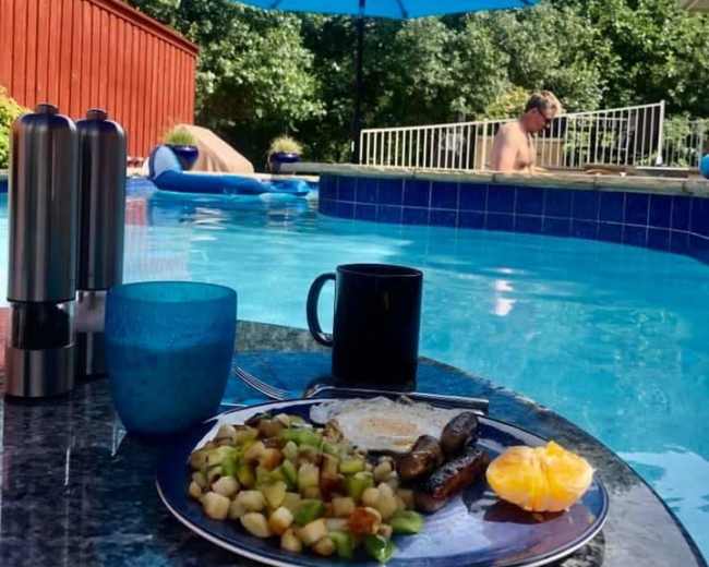 A plate of breakfast with fruit, sausages, and an egg sits on a table beside a swimming pool, with a blue umbrella providing shade, while two people relax in the water.