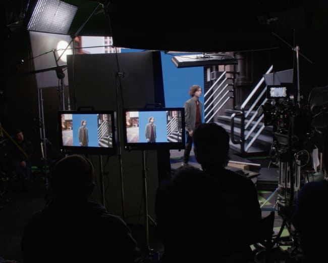 A person walks on a set with two monitors displaying their image, surrounded by film equipment and crew members in the foreground.