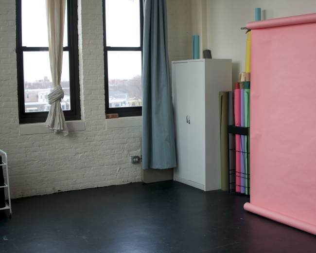 The image shows a studio space with a pink backdrop on a stand, a cabinet, and rolls of colorful paper stored nearby.