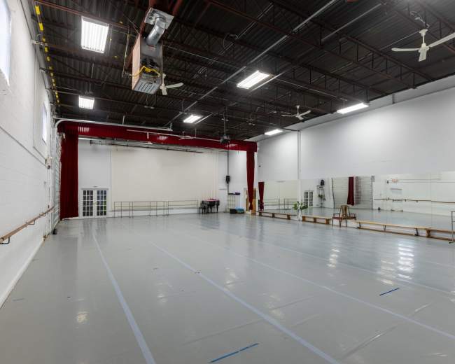 The image shows a spacious dance studio with a mirrored wall, wooden ballet barres, and a polished floor under bright overhead lights.