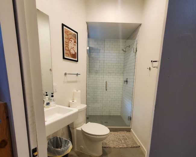 The image shows a bathroom featuring a shower, toilet, sink, and a small mirror, with gray concrete flooring.