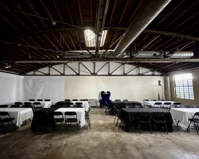 The image shows an empty event space with rectangular tables covered in black and white tablecloths arranged in a symmetrical layout and a large blue chair in the background.